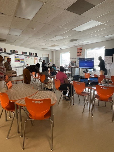A teacher presents at the front of a classroom while participants sit at tables with laptops, following along with a presentation on a screen.