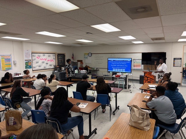 Adults sit at classroom desks facing a teacher presenting on a screen, engaged in a professional development session.