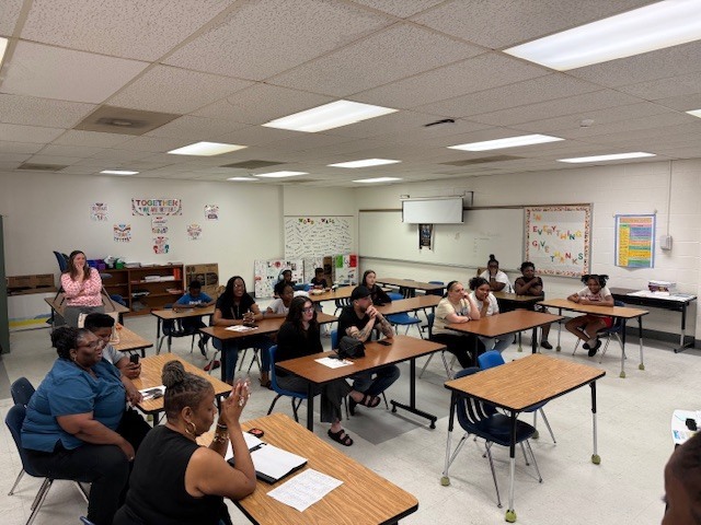 Group of adults seated at desks in a classroom, smiling and clapping during a training or meeting.