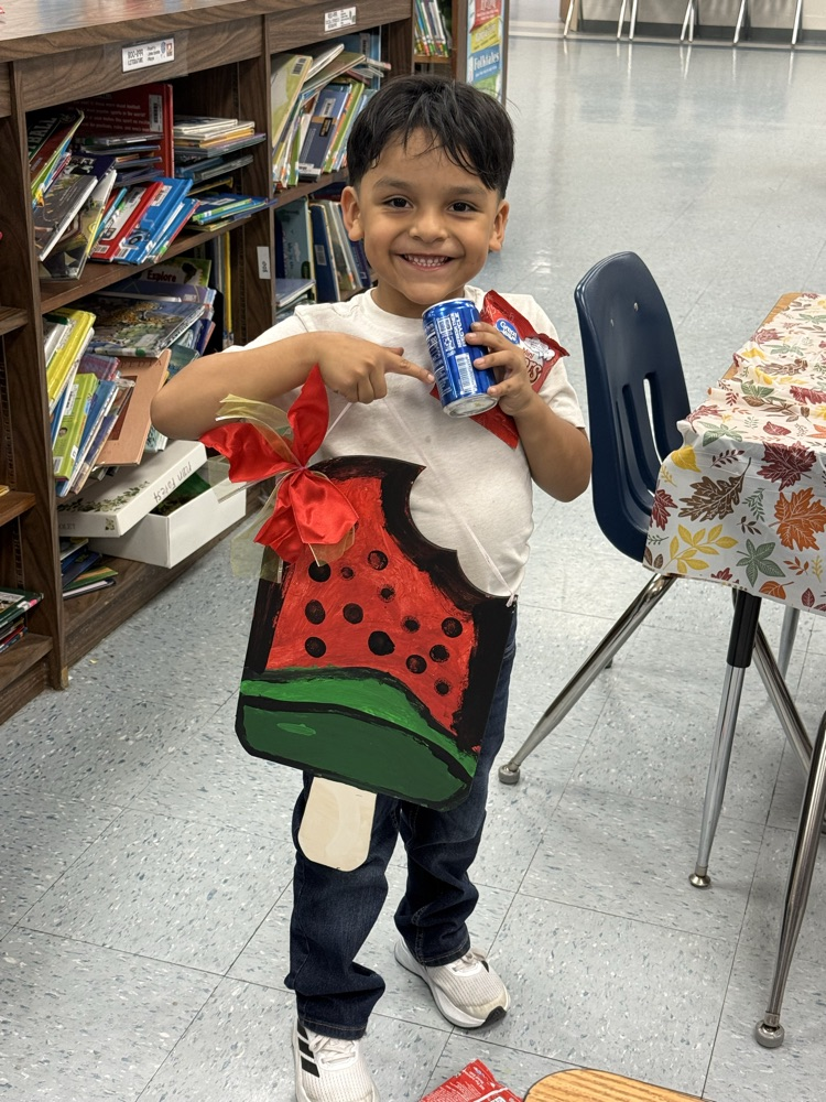 A smiling young boy stands in a classroom holding a painted watermelon popsicle sign with a bite taken out of it and a red bow. He holds a drink and snack, proudly showing his artwork.