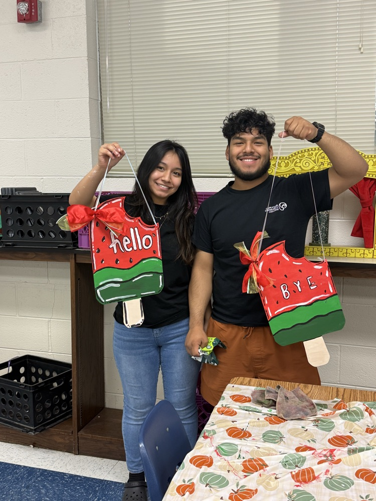 Two teens stand side by side holding painted watermelon popsicle signs. One sign says “hello” and the other says “bye,” both decorated with bright colors and bows.