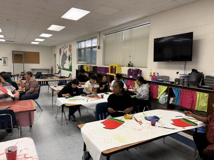 Three participants sit at a table in a classroom working on watermelon-themed paint projects, with paint supplies, brushes, and palettes spread out in front of them.