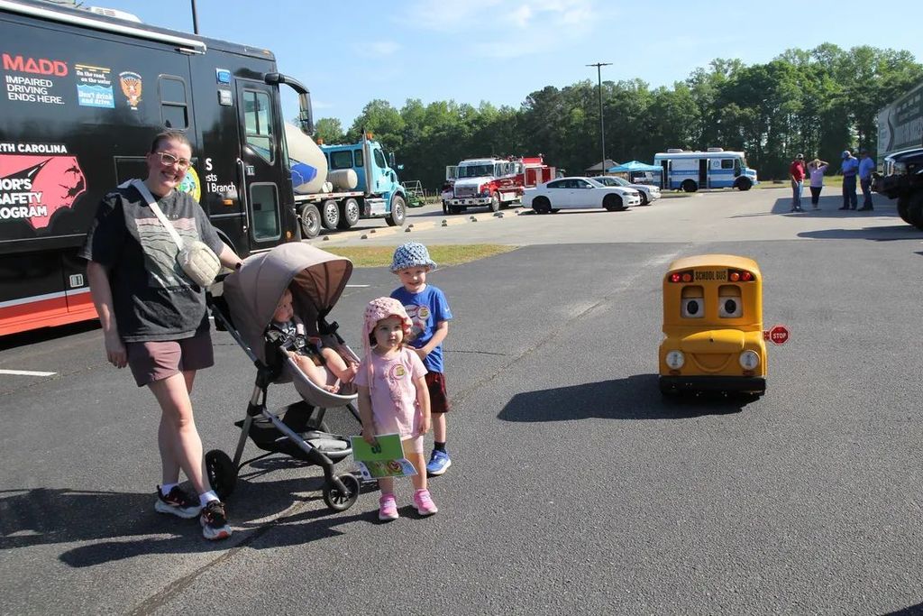 A mom with three small children, one in a stroller stand next to a small school school bus.  You can see a firetruck and cement mixer in the background.