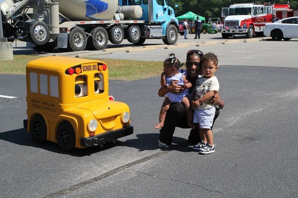 A parent and two small children are crouched beside a small school bus. You can see a fire truck and a cement mixer in the background.