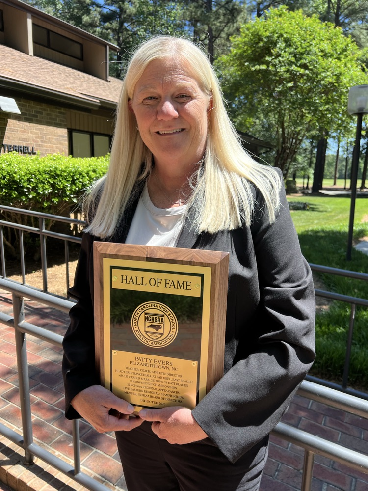 coach Patty Evers in a black suit holding her Hall of Fame plaque. It is a sunny day and there’s a brick path behind her and some green grass.