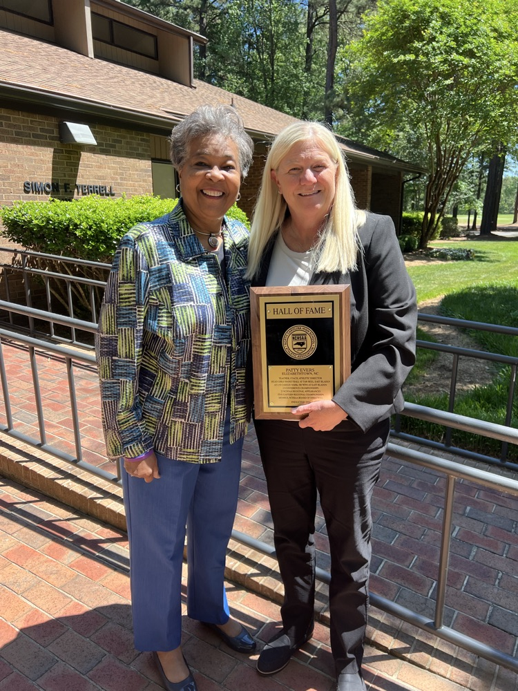 COACH PATTY EVERS IN A BLACK SUIT WITH HER HALL OF FAME PLAQUE AND A LADY DRESSED IN BLUE PANTS