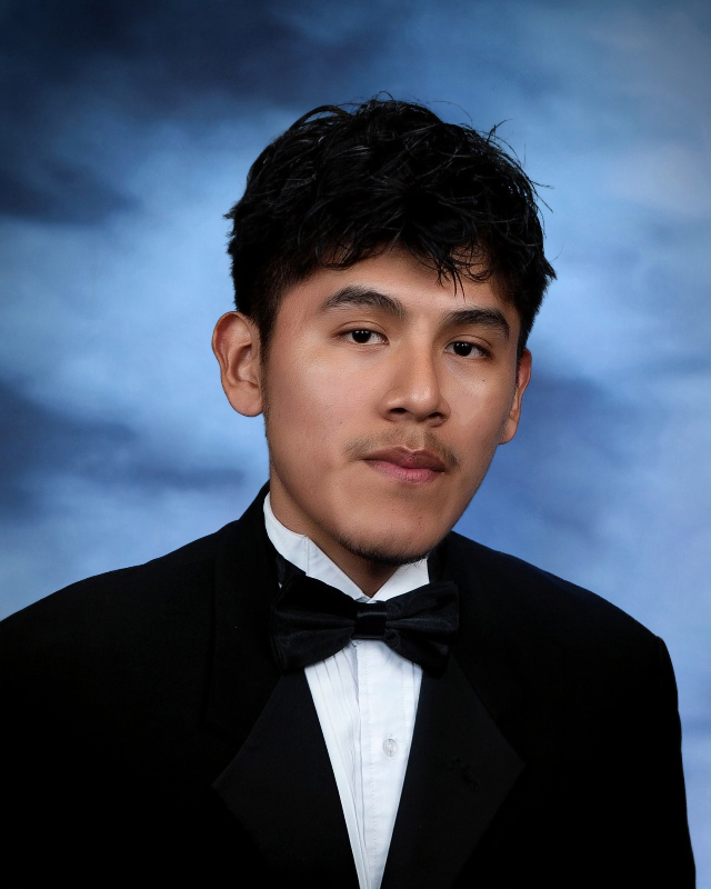 Michael Reyes Maldonado, a high school senior, wearing a black tuxedo and bow tie, poses for a formal studio portrait against a blue cloudy backdrop, looking directly at the camera with a neutral expression.