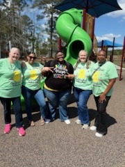 Staff members in matching shirts pose together, smiling. The sky is bright blue with scattered clouds, creating a cheerful outdoor setting.