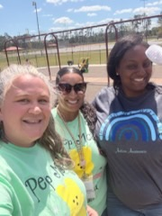 Staff members in matching shirts pose together, smiling. The sky is bright blue with scattered clouds, creating a cheerful outdoor setting.