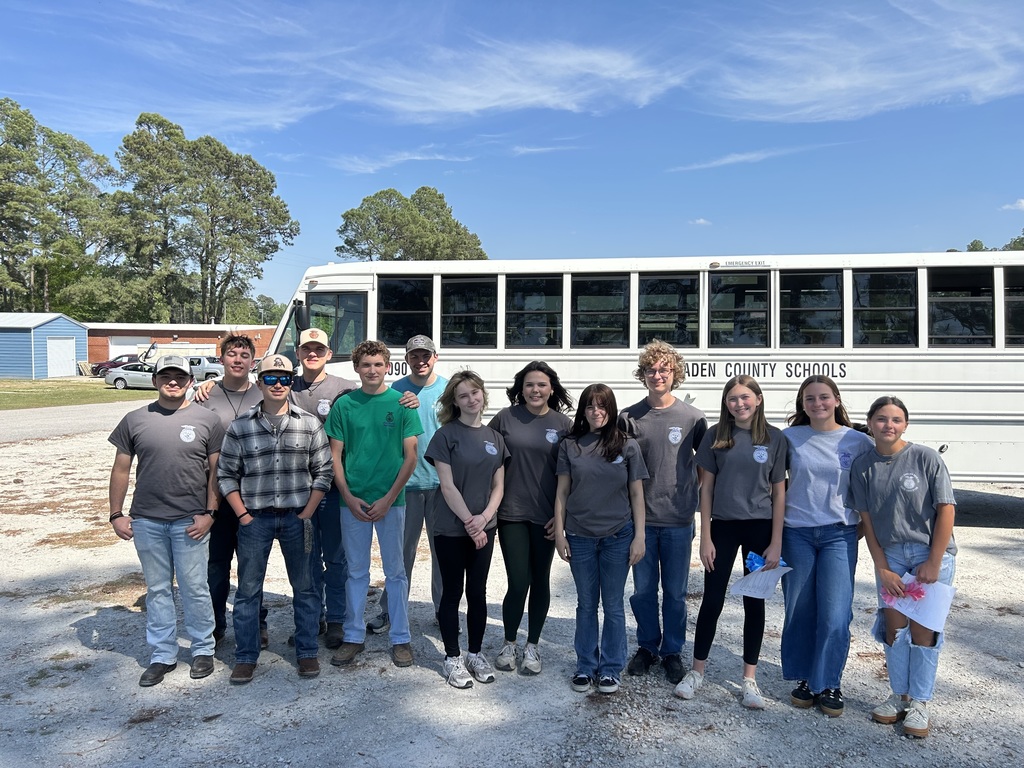 A group of thirteen students poses for a group photo in front of a white school bus labeled "Bladen County Schools." They are outdoors on a gravel lot under a clear blue sky. Most students are wearing matching grey t-shirts with a circular logo, while others are in green or plaid shirts.