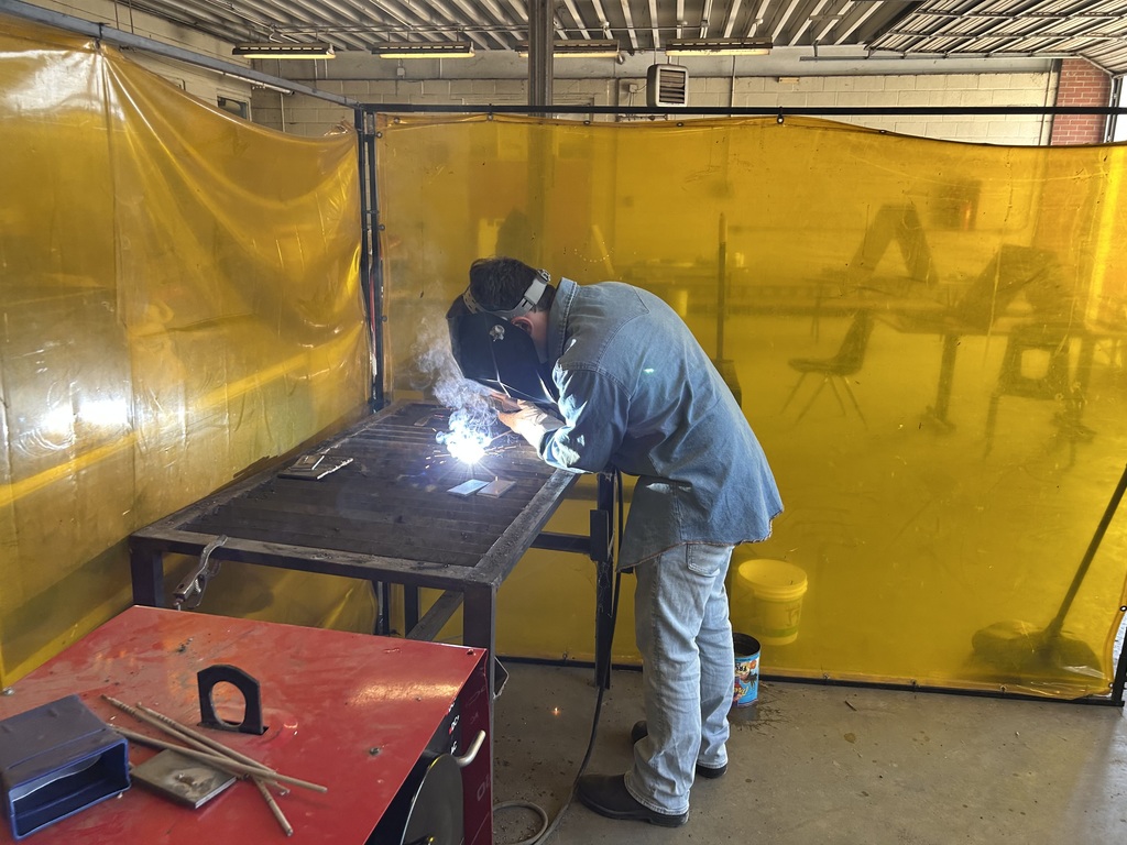 A student in a denim work shirt and welding helmet performs a weld on a metal table, creating a bright blue arc of light and smoke. The workspace is enclosed by yellow semi-transparent safety curtains in a large industrial shop.