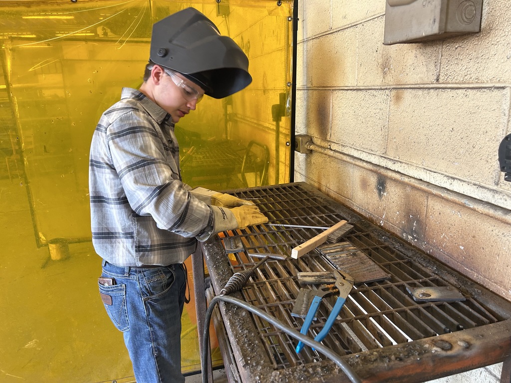 A student wearing a plaid shirt, denim jeans, work gloves, and a welding helmet prepares a metal workpiece on a slatted metal table. A yellow translucent welding curtain hangs in the background, and various metal-working tools, such as pliers and a wire brush, sit on the table.