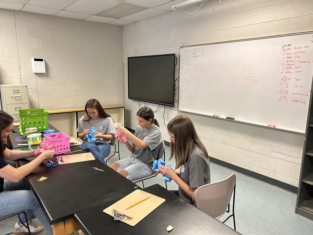 Three female students sit at a black lab table in a classroom, carefully tying blue and pink ribbons into decorative bows. A teacher or instructor assists from the left side of the table. In the background, a large digital screen and a whiteboard with handwritten technical notes are visible against a white cinderblock wall.
