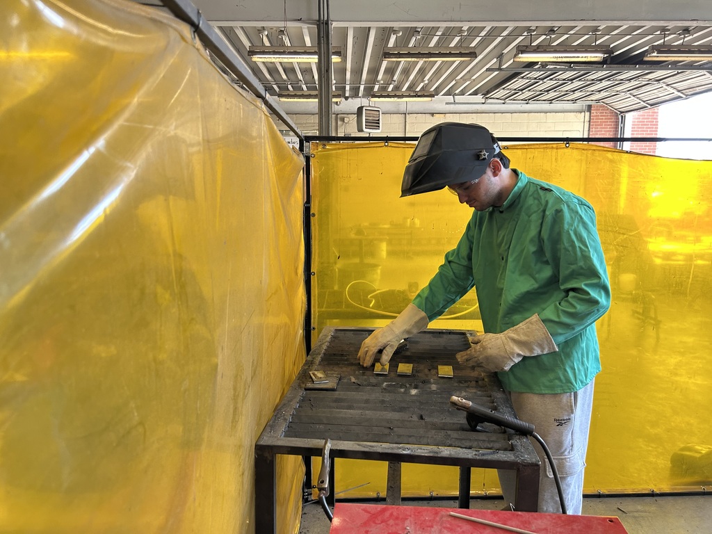 A student wearing a green welding jacket, grey joggers, and a welding helmet stands behind a yellow safety curtain, adjusting metal plates on a slatted welding table in preparation for a project.
