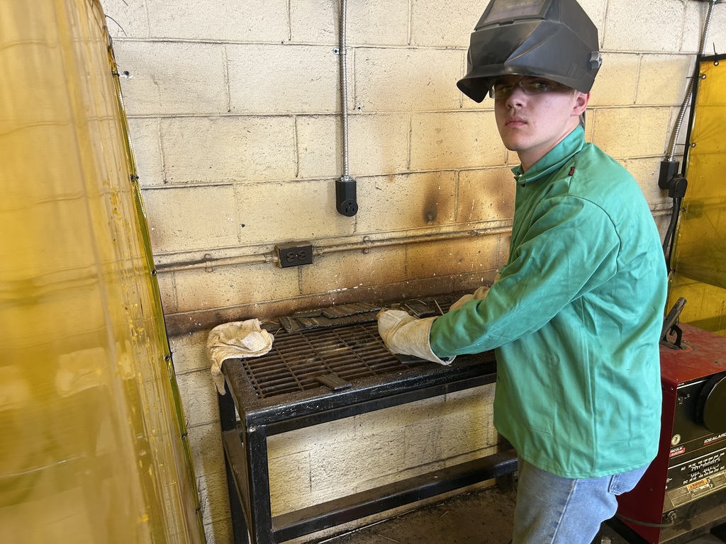 A student in a bright green protective welding jacket and a welding helmet stands at a metal workbench in a shop. He is wearing heavy-duty gloves and looking toward the camera. The wall behind him is made of tan cinderblocks with electrical conduits and outlets.