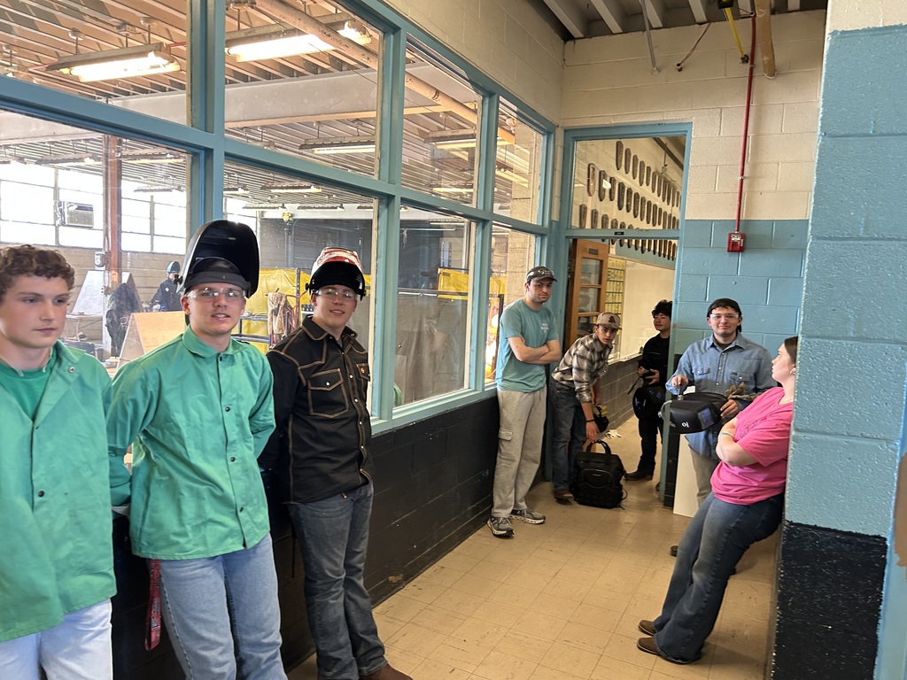 A group of students stands in a hallway outside a welding workshop. Several students wear protective welding jackets in green and black, and two have welding helmets flipped up on their heads. They are looking toward the camera, and large glass windows behind them provide a view into the industrial shop area.