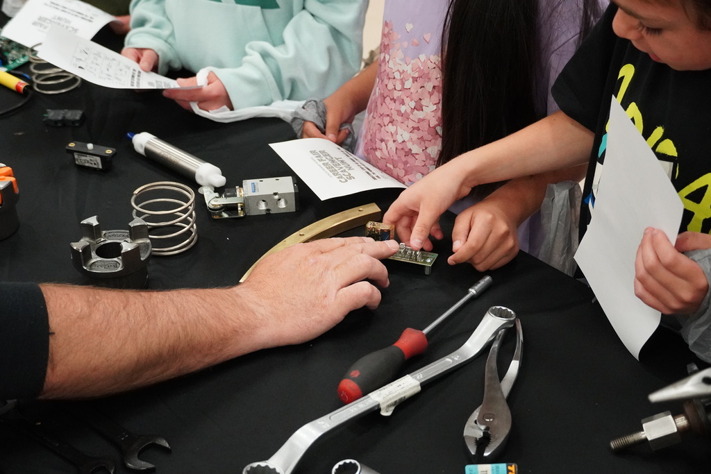 And adult is pointing out a tool to a student. The table is full of different types of tools.