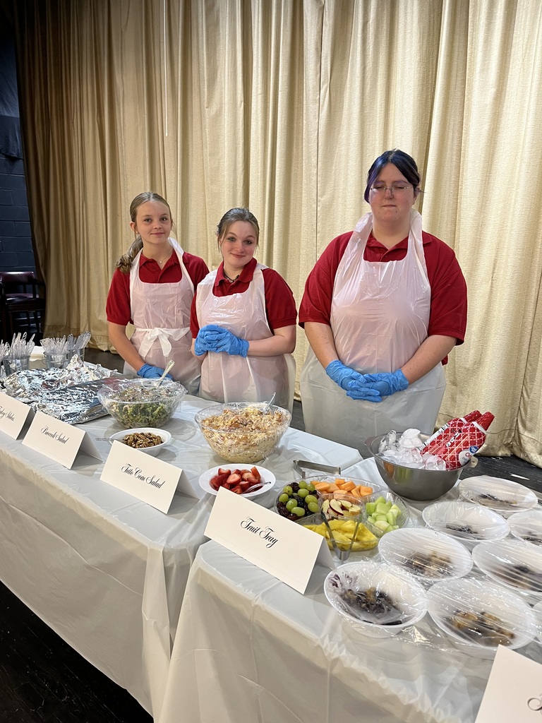3 students in white plastic aprons posing in front of a serving table