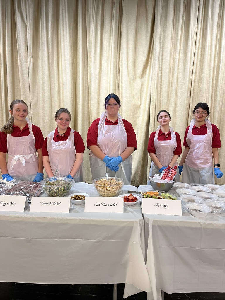 A group of five female students in matching aprons and hairnets stand behind a buffet table of food during a culinary event, smiling and posing for a photo in a bright room