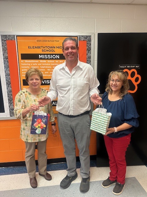 Three staff members stand together smiling in a school hallway, each holding a small gift bag. A bulletin board behind them displays Elizabethtown Middle School’s mission statement in orange and black school colors.