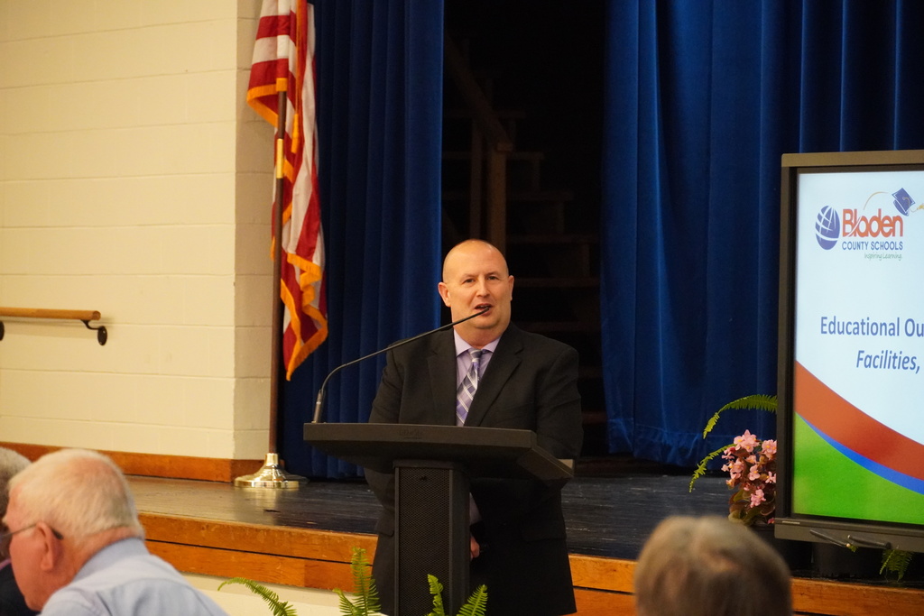 An adult man in a suit is speaking from a podium. There is an American flag behind him and you can see part of a slide deck on a large screen. 