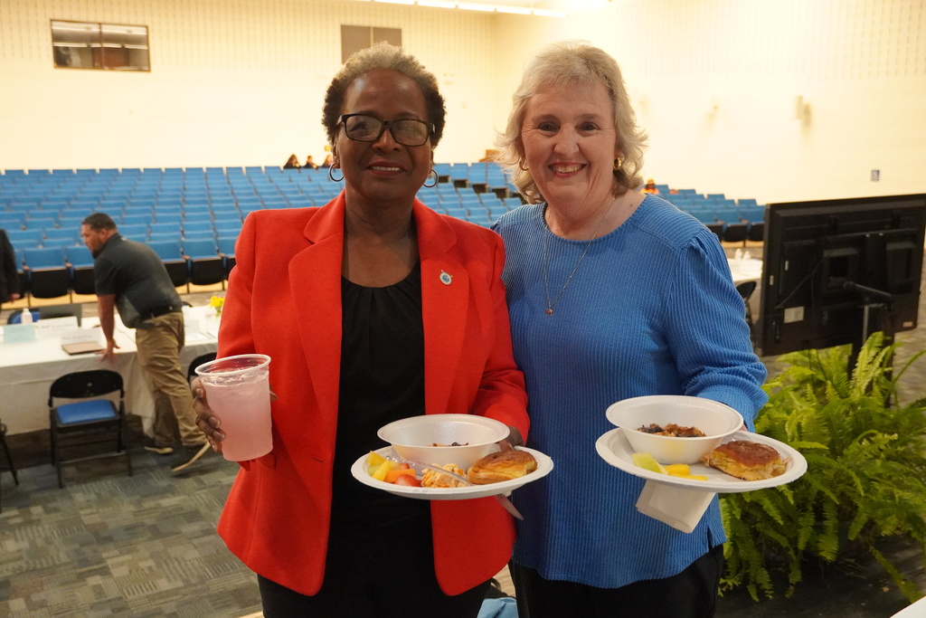 Two women stand together holding plates of food. In the background you can see a large monitor, ferns, and blue auditorium seats.