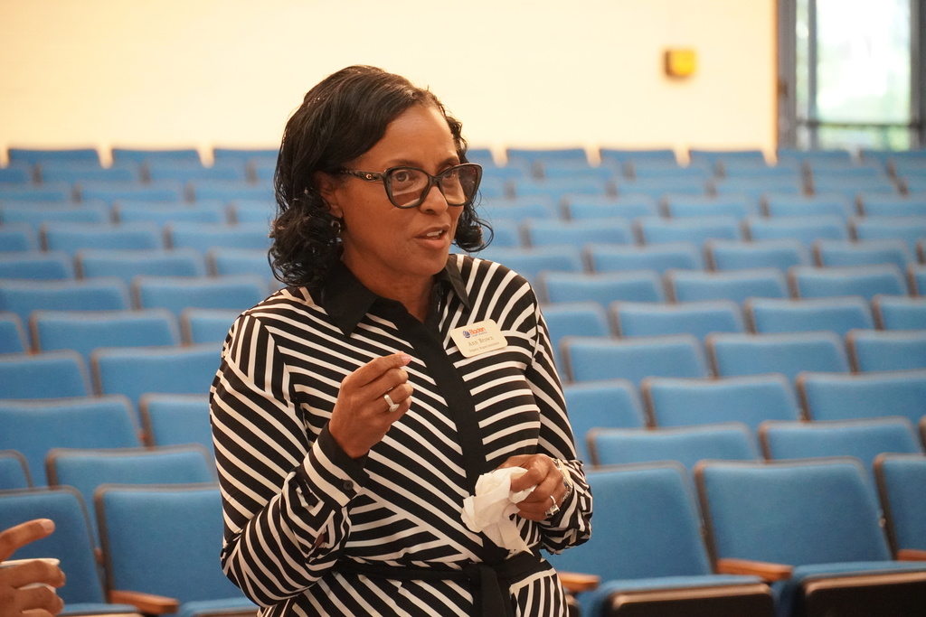 A woman in a black and white striped dress is speaking. You can see blue auditorium seats behind her.