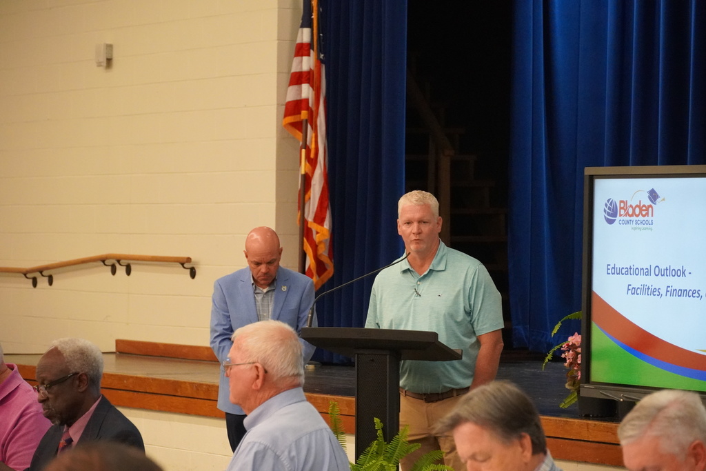 Two adults stand at a podium addressing a group of people. You can see the American flag behind them and part of a slide deck on a large monitor.