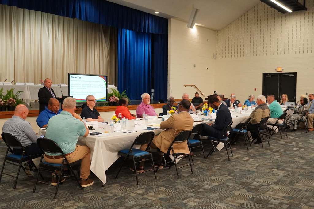 A group of adults sit at a long table in an auditorium listening to a gentleman speak at a podium.