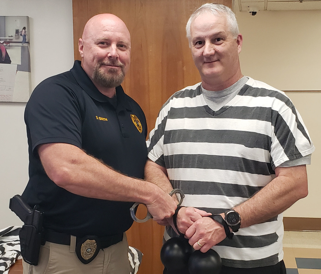 Two men pose indoors, one dressed as a police officer placing handcuffs on another man wearing a striped shirt, holding black balloons, smiling during a Jail-a-thon fundraiser event.