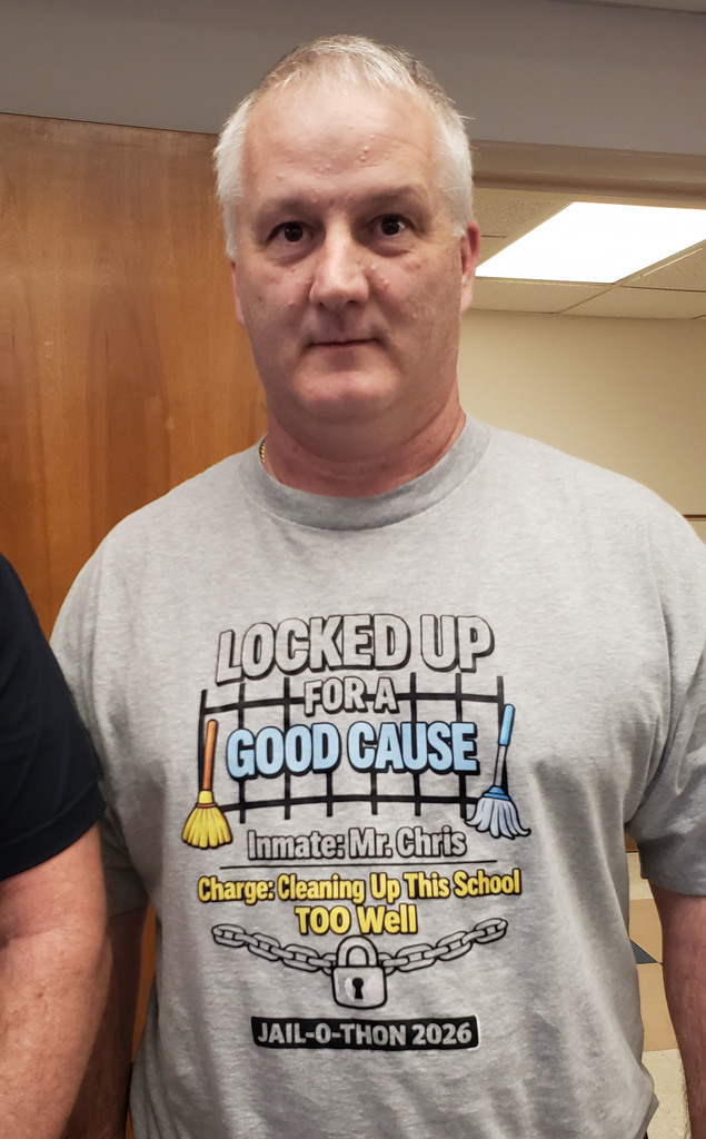 A man with short gray hair wearing a gray “Locked Up for a Good Cause” Jail-a-thon 2026 T-shirt stands indoors in front of a wooden door.
