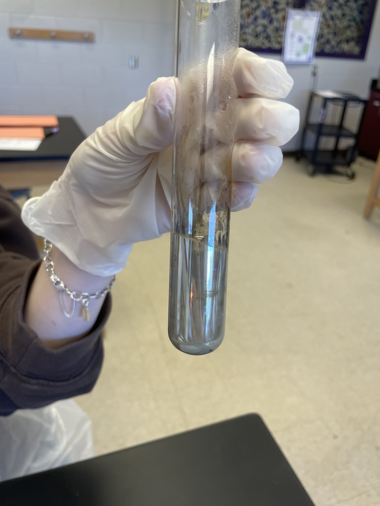 A gloved hand holds a glass test tube containing a silver, reflective liquid. The mirror-like coating on the glass reflects the surrounding chemistry lab environment.