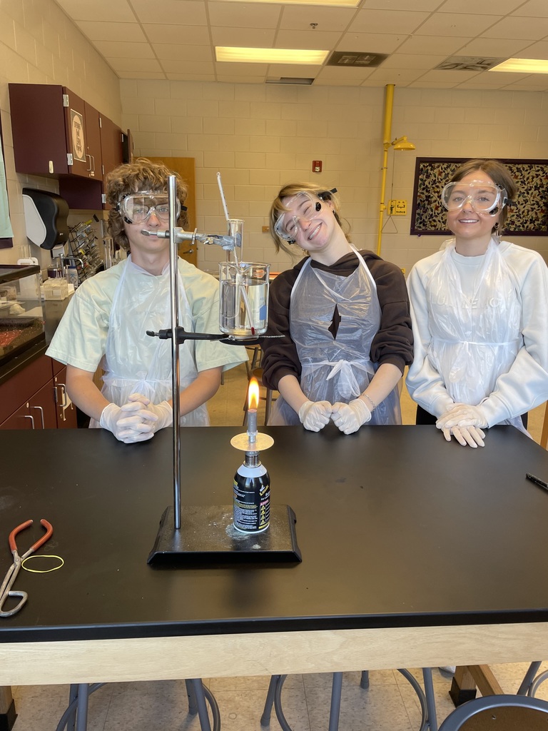 Three Honors Chemistry students in lab coats and gloves stand behind a lab bench, observing a lit Bunsen burner during a silver mirror redox reaction experiment