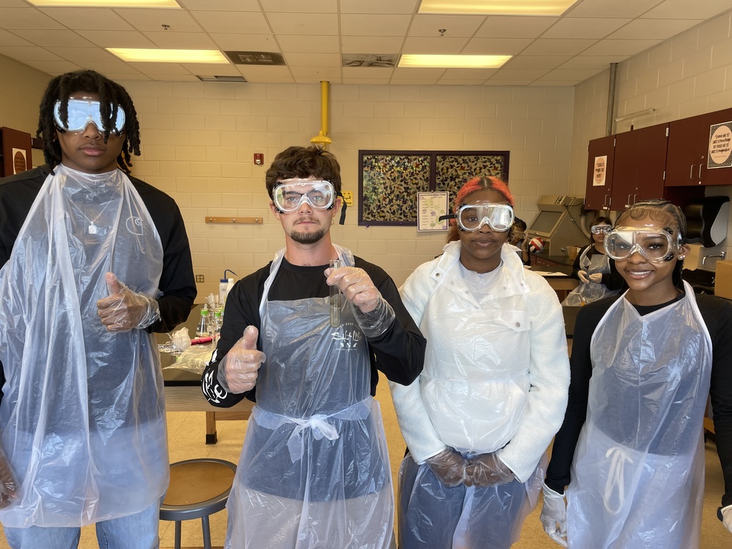 Four West Bladen High chemistry students in safety goggles and aprons smile in a lab. One student holds a test tube containing a silver mirror finish from a successful Tollens' test.