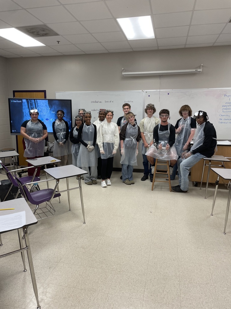 A group of high school chemistry students in lab aprons and safety goggles pose together in a classroom. They are standing in front of a whiteboard and a digital screen showing lab notes