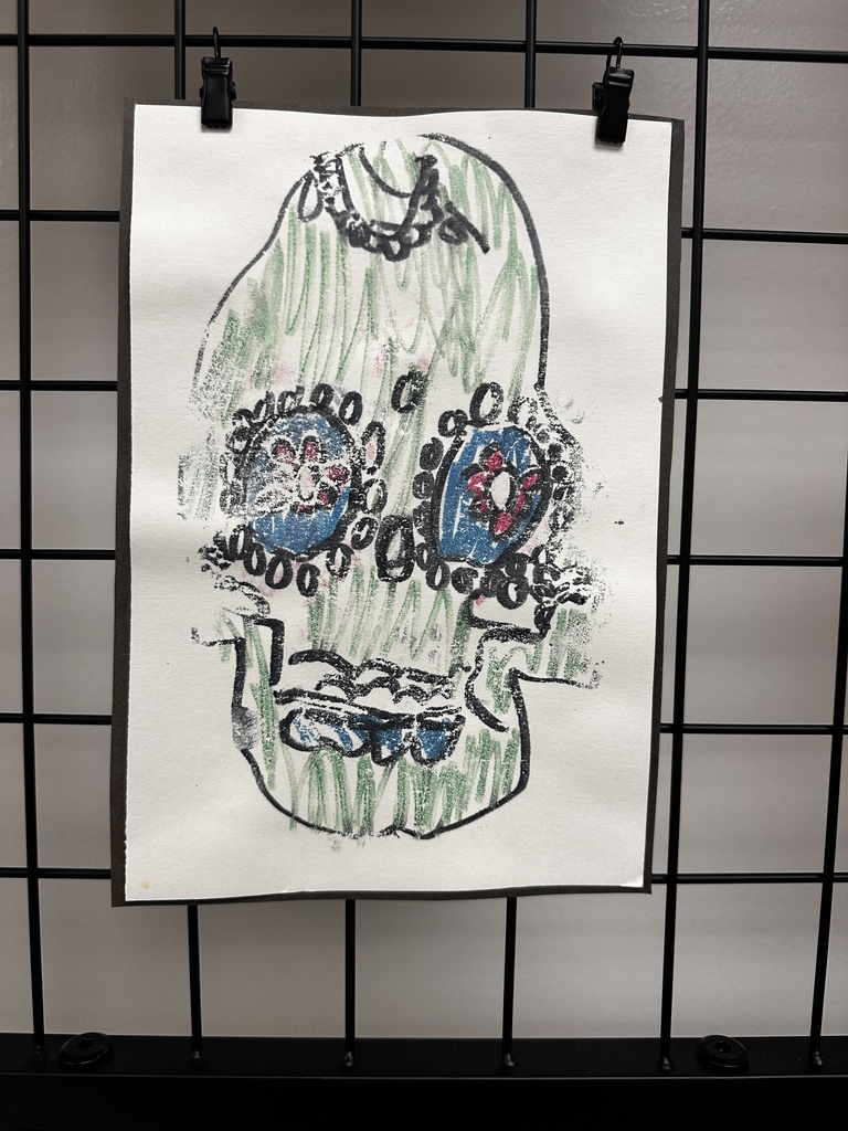 Drawing of a decorative sugar skull with black outline, green crayon-filled interior, ornate circular eyes with blue centers containing red and white flower patterns surrounded by black dots, a small heart-shaped nose with blue coloring, wavy lines for the mouth, and a decorative black chain or garland at the top.