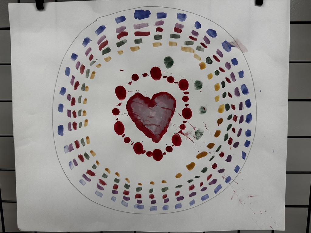 Colorful painting on white paper featuring a large red heart surrounded by an inner ring of red and green dots with red paint splatters, enclosed by a light gray circle, and decorated with concentric arcs of small colorful brush strokes and stamps in blue, purple, red, green, yellow, and orange creating a mandala-like pattern.