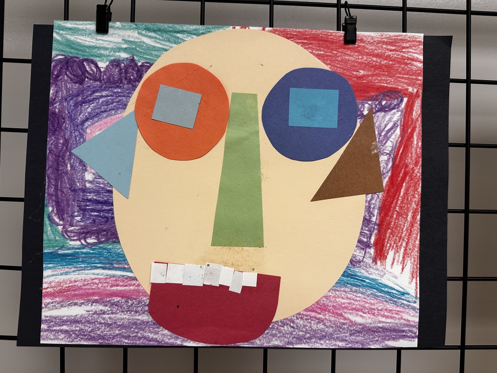 Child's mixed-media collage and crayon artwork featuring a large tan oval face with colored paper circle eyes (orange with light blue rectangle on left, dark blue with bright blue rectangle on right), a green trapezoidal nose, and a red semi-circular mouth with white paper teeth. The background includes purple, teal, pink, and red crayon scribbles with light blue and brown paper triangles on either side.