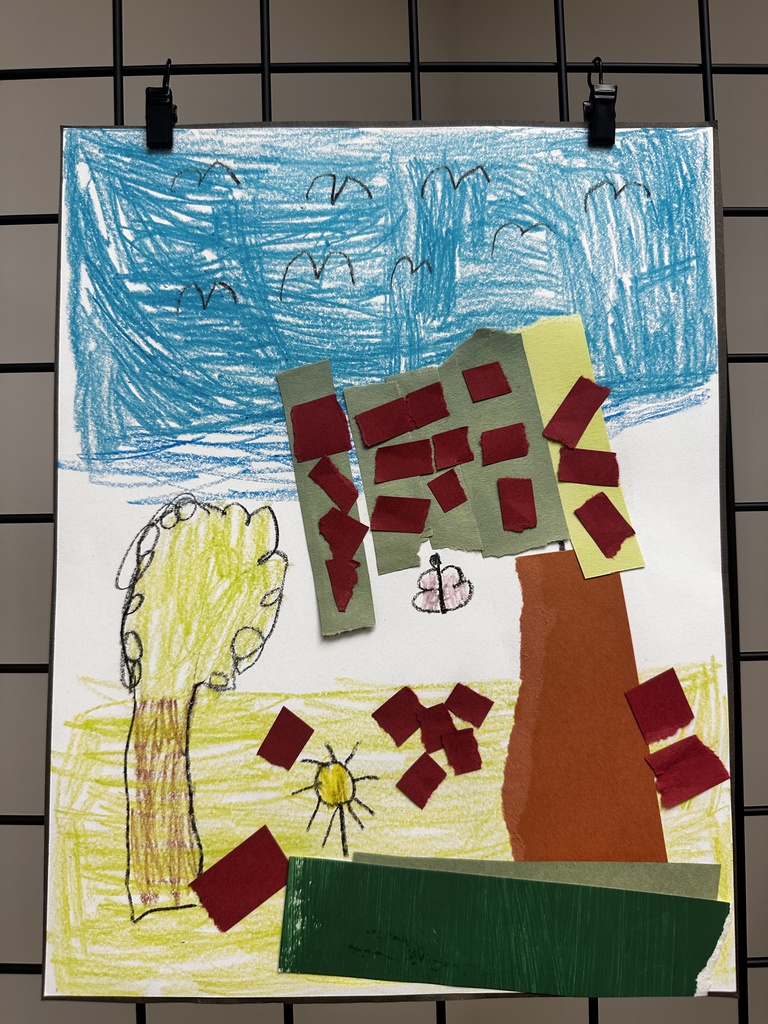 Child's mixed-media artwork combining drawing and collage showing an outdoor scene with blue scribbled sky and black bird shapes, yellow ground, a tree with brown trunk and green leaves on the left, a yellow sun with black rays in the center, and collaged paper buildings in green and yellow with red windows. Red paper rectangles are scattered across the ground, and a small drawn butterfly appears in the middle area.