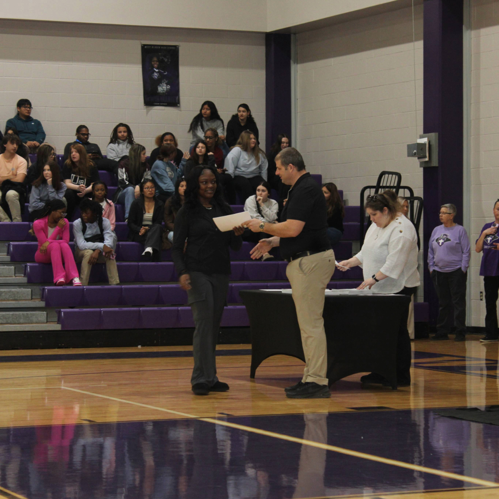 Mr. Orrock, in a black shirt and khakis hands an award certificate to a smiling female student on a gym floor. Other students sit in bleachers in the background during the ceremony
