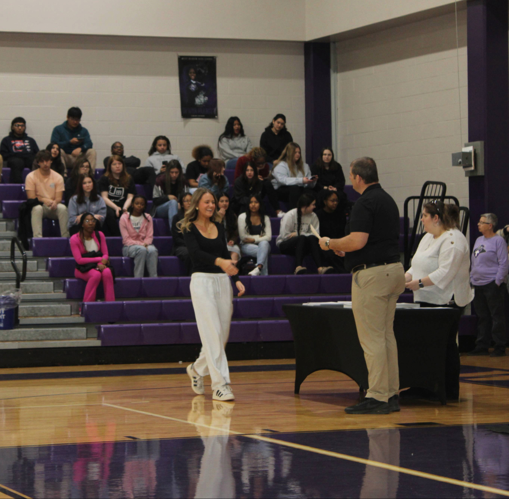 Mr. Orrock in a black shirt hands an award certificate to a smiling female student in a gymnasium. Other students watch from purple bleachers during the academic ceremony.