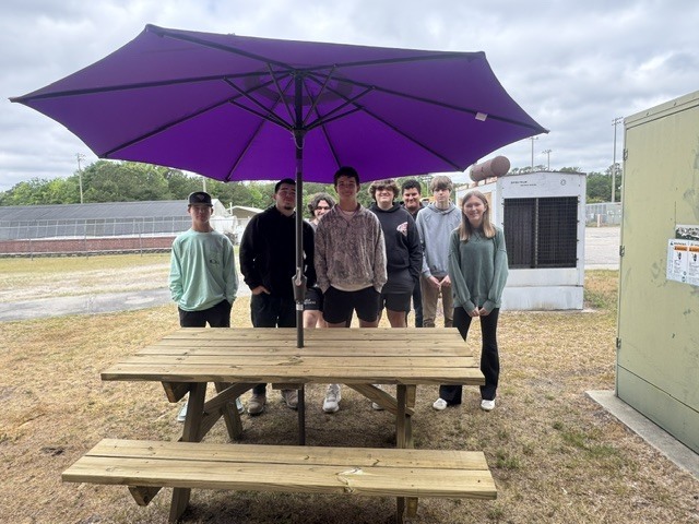 A group of students stands and sits around a newly built wooden picnic table outdoors. A large purple umbrella is mounted in the center of the table, open above them. Three students sit at the table in the foreground, while several others stand behind it, smiling or looking toward the camera. The setting appears to be a school campus with grass, a greenhouse structure, and utility buildings visible in the background under a cloudy sky.