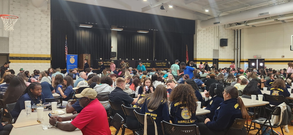 A large group of students and adults sit at tables in a gymnasium for an event. Banners are visible in the background, and a person in a red shirt is in the foreground.