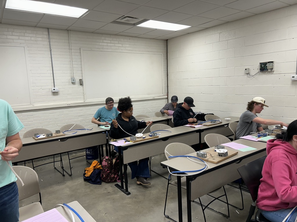 Students sit at classroom tables working on hands-on projects with blue and white wires. An instructor stands at the front of the room near a whiteboard in a bright, educational setting