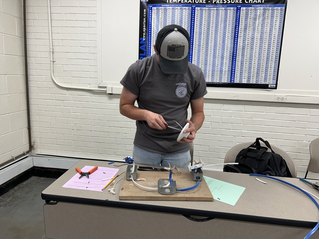 A student in a grey West Bladen FFA shirt and cap works on a mechanical project with wires and tools at a desk. An agricultural temperature and pressure chart hangs on the wall behind him