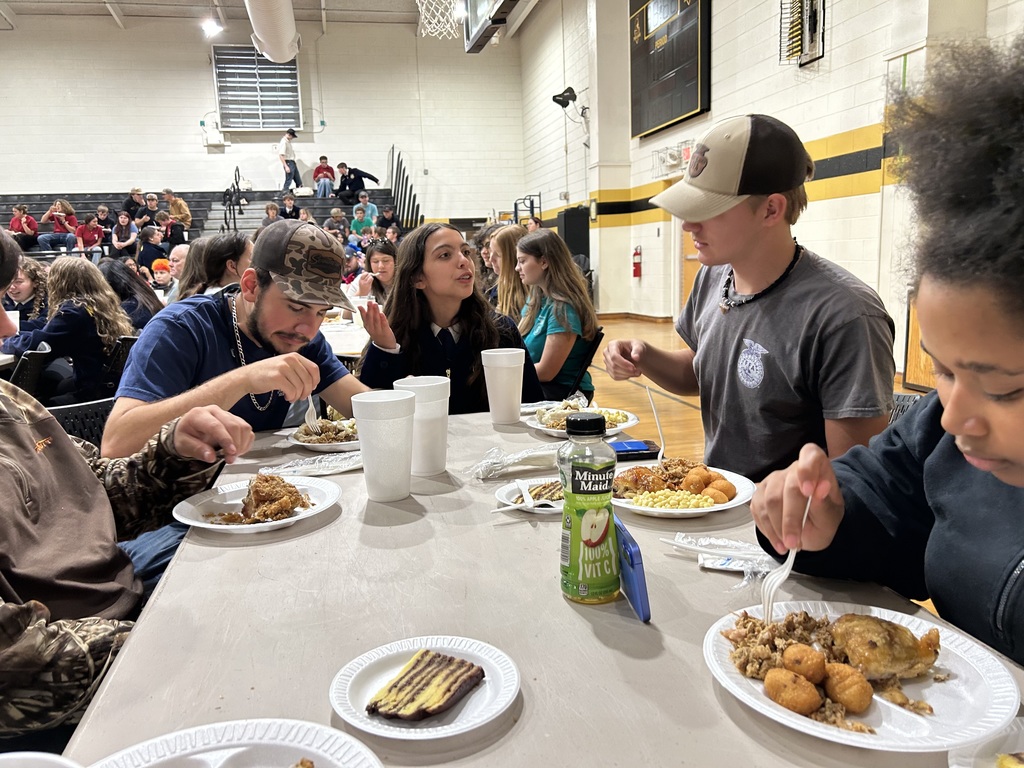 A group of high school students sit at a long table in a gymnasium, eating a meal together. A student in a grey shirt and trucker hat focuses on his plate in the foreground.