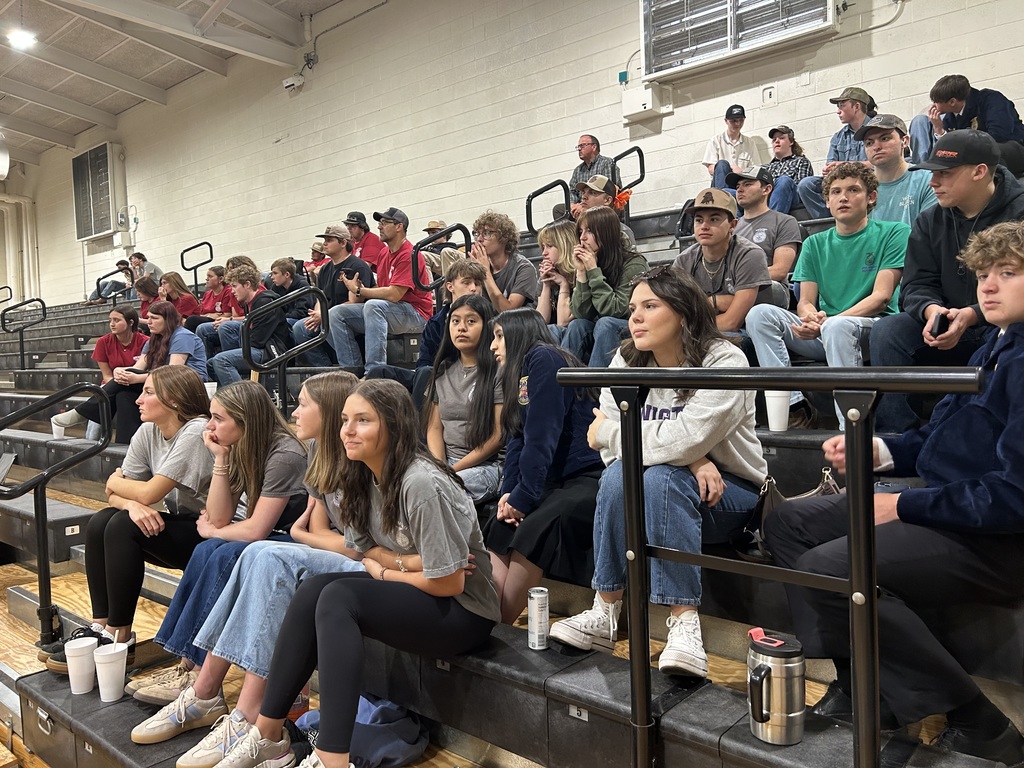 A group of students sits on bleachers in a gymnasium, looking toward a presentation. Several students in the front wear FFA blue and gold jackets, while others are in casual attire.