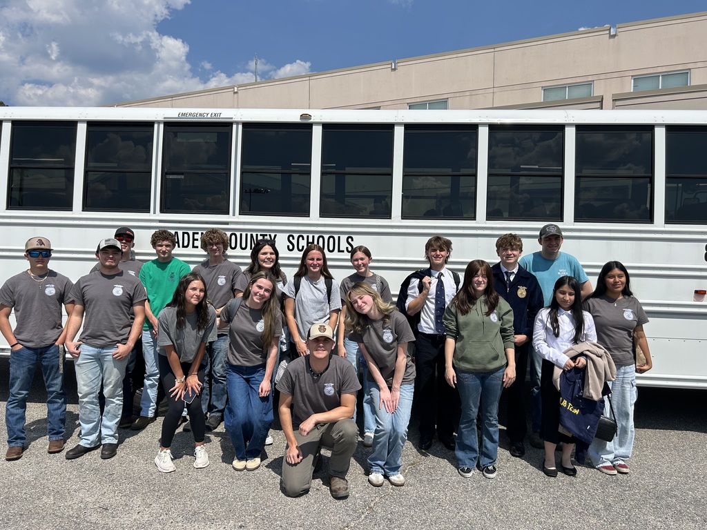 A large group of West Bladen FFA students and advisors pose for a group photo in front of a white school bus on a sunny day. They are wearing matching grey and green FFA shirts.