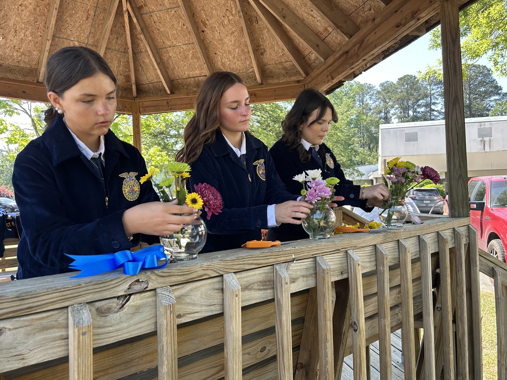 Three female students in navy blue FFA jackets carefully arrange vibrant sunflowers and greenery into glass vases on a wooden railing during an outdoor floral competition