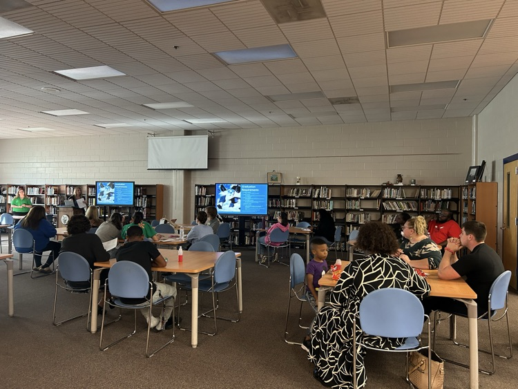 several people sitting at tables in a library looking at two screens while a woman presents information